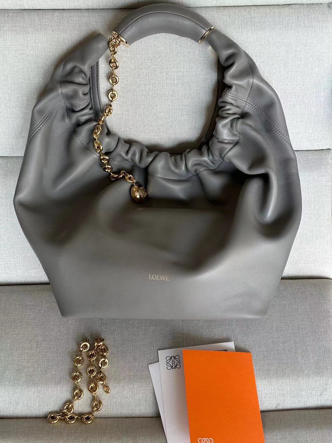 LOEWE Small Squeeze Bag: Where Elegance Meets Innovation in Luxurious Nappa Leather. The Squeeze bag embodies sophistication and femininity, crafted from luxurious nappa leather. It stands as a masterpiece, seamlessly blending traditional craftsmanship, innovative research, and distinctive design elements. The bag features a surprisingly lightweight ruched body, complemented by an adjustable donut chain and a tactile, squeezable handle. This particular version showcases the elegance of nappa lambskin in a compact size, adding an extra touch of refinement to its allure.