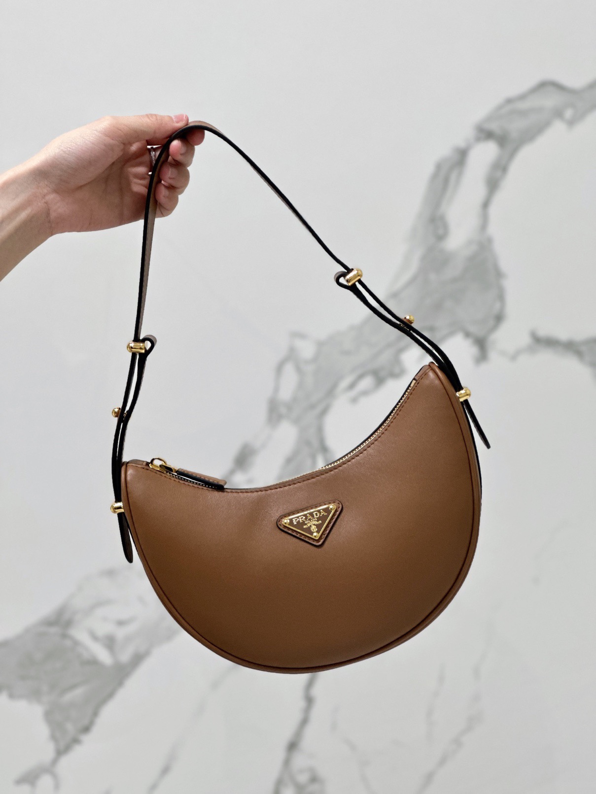 PRADA Leather Shoulder Bag. High-End Quality Bag including gift box, care book, dust bag, authenticity card. This sleek leather bag expresses versatile, modernist allure. The accessory with a curvy silhouette is completed by the leather extension for versatile wear. With a nappa leather lining and metal hardware, the bag is accented with the iconic enameled metal triangle logo on the front.