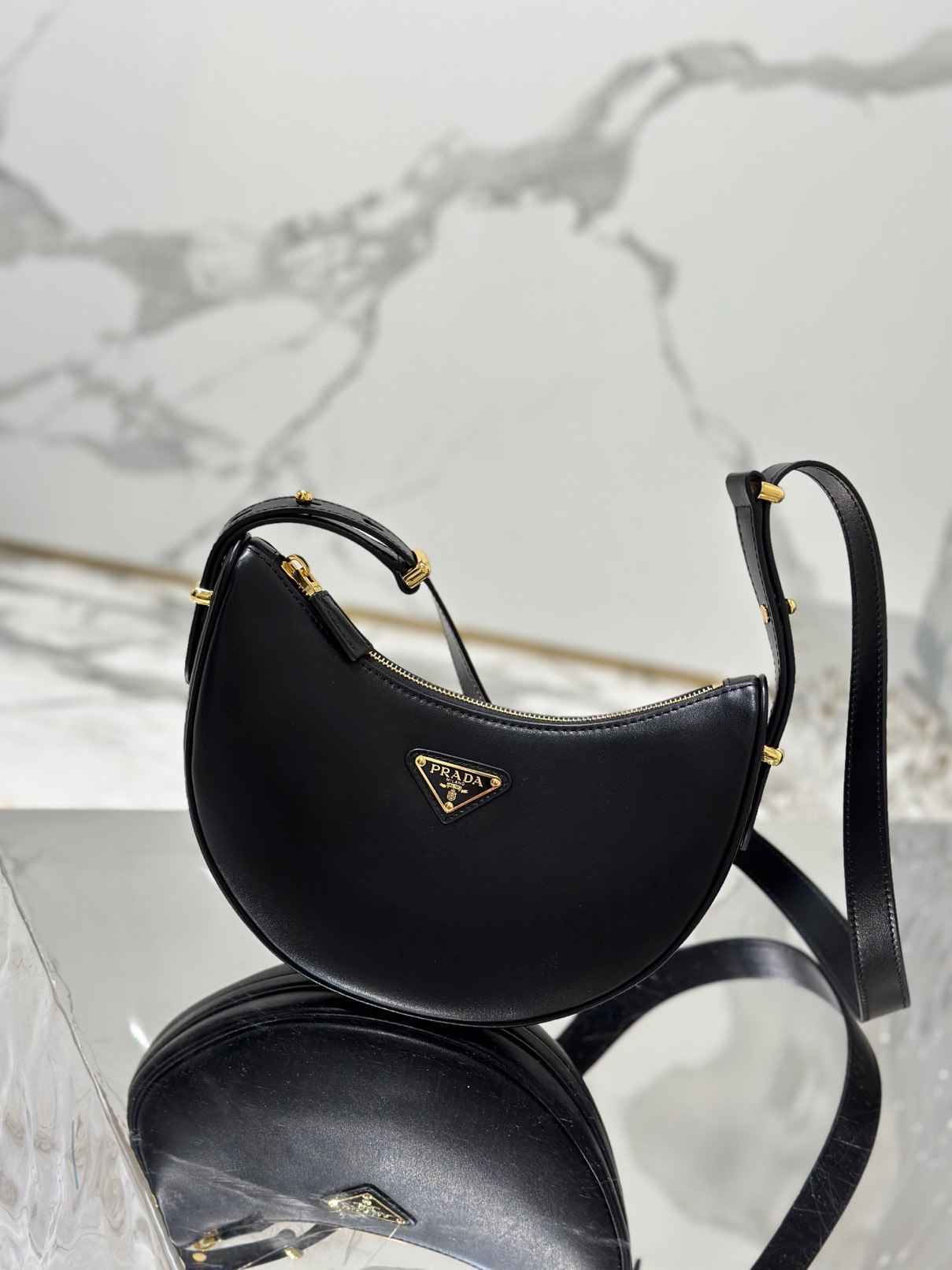 PRADA Leather Shoulder Bag. High-End Quality Bag including gift box, care book, dust bag, authenticity card. This sleek leather bag expresses versatile, modernist allure. The accessory with a curvy silhouette is completed by the leather extension for versatile wear. With a nappa leather lining and metal hardware, the bag is accented with the iconic enameled metal triangle logo on the front.